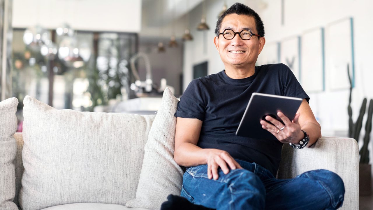 Mature aged Asian man sitting on sofa with a digial tablet in hand and smiling