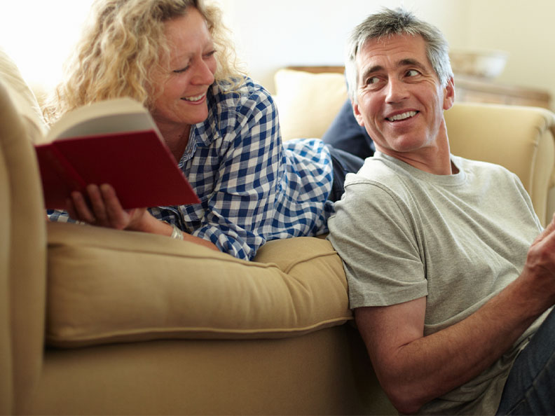 A couple looking at eachother as they read books together in their lounge room on their couch in their home. A man is using a tablet and a woman is holding a hardcover book.
