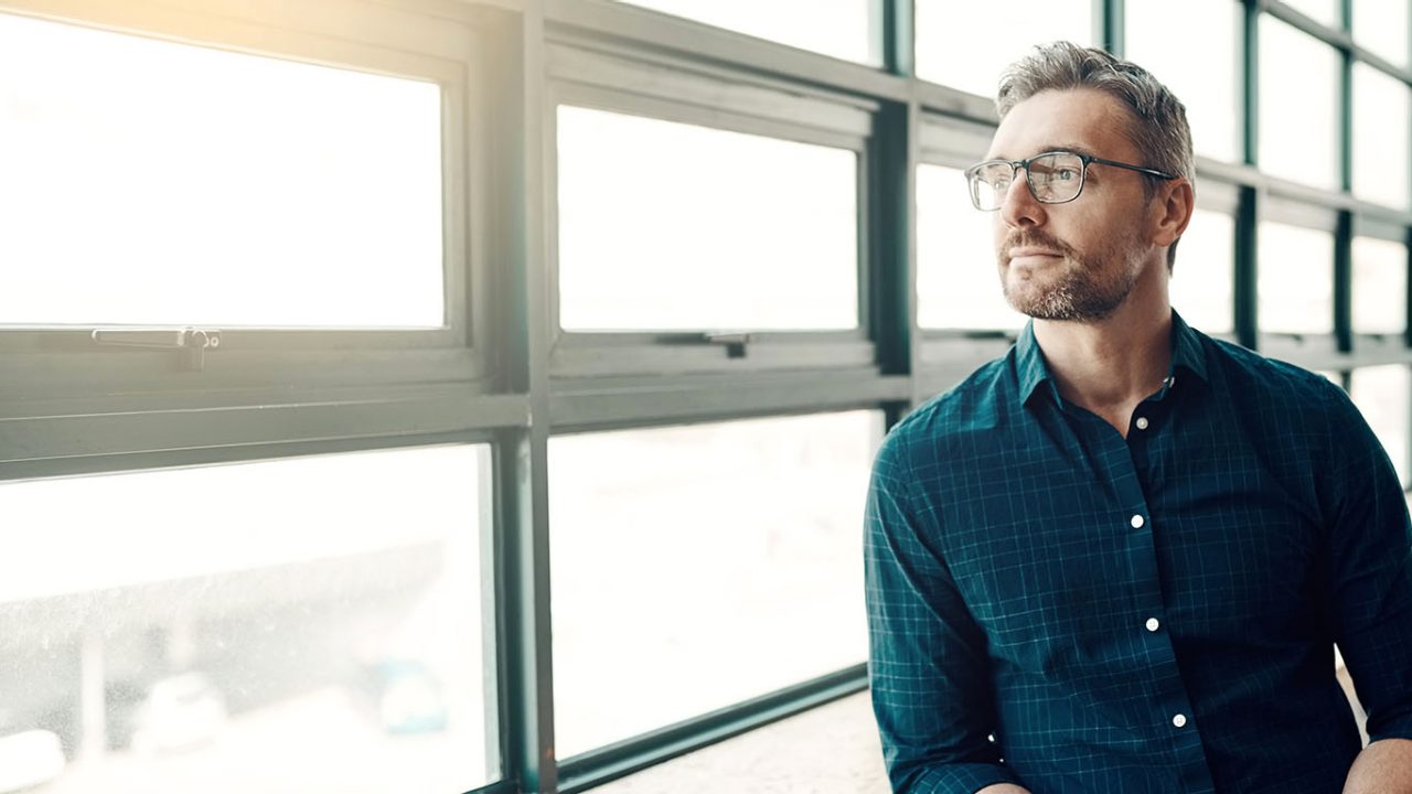 A middle aged man with glasses in smart casual clothing looking out a window of a modern office