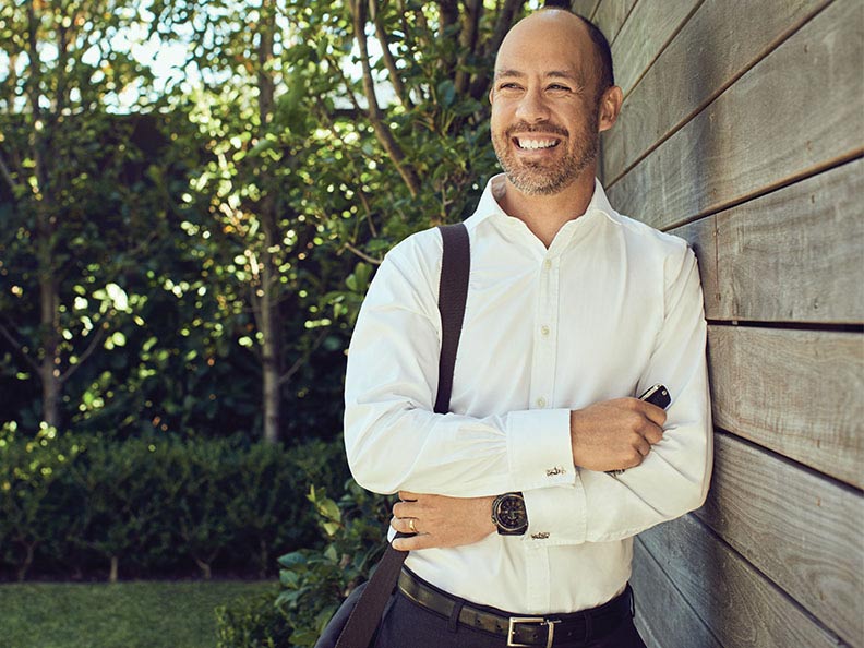 A man dressed in a white dress shirt with a bag strap on his right shoulder smiling and leaning outside against a wall.