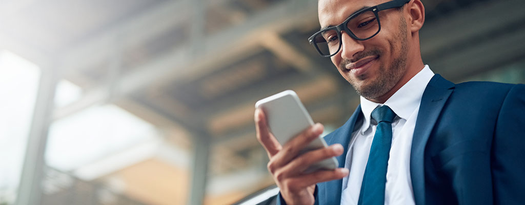 Shot of a young businessman using his phone while standing outside leaning against a car. He’s wearing a suit.