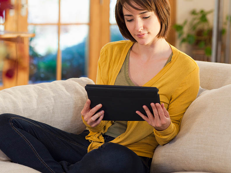 A woman with brown hair and wearign a yellow cardigan top sitting in a warmly lit lounge holding and reading a digital tablet with two hands