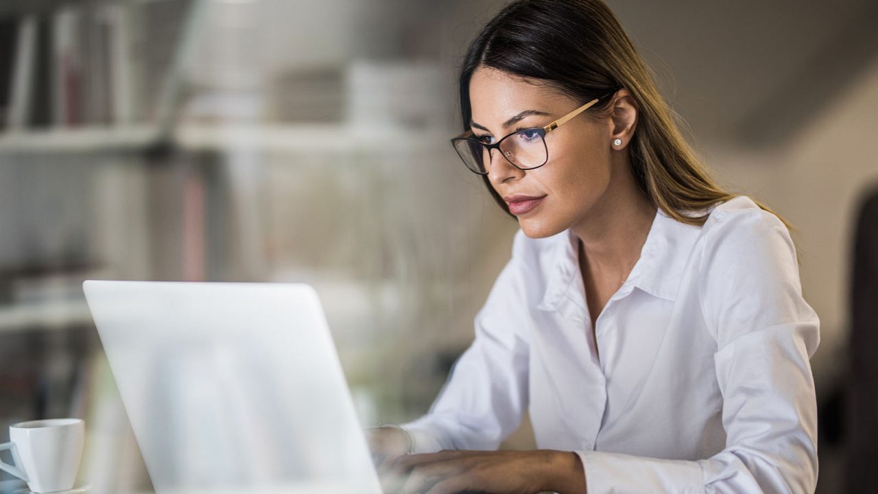 A mid-shot of a young business woman with glasses on working on a laptop in a quiet looking office.