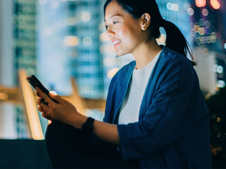 A young woman sitting outside at night looking at a mobile phone with city lights in the background