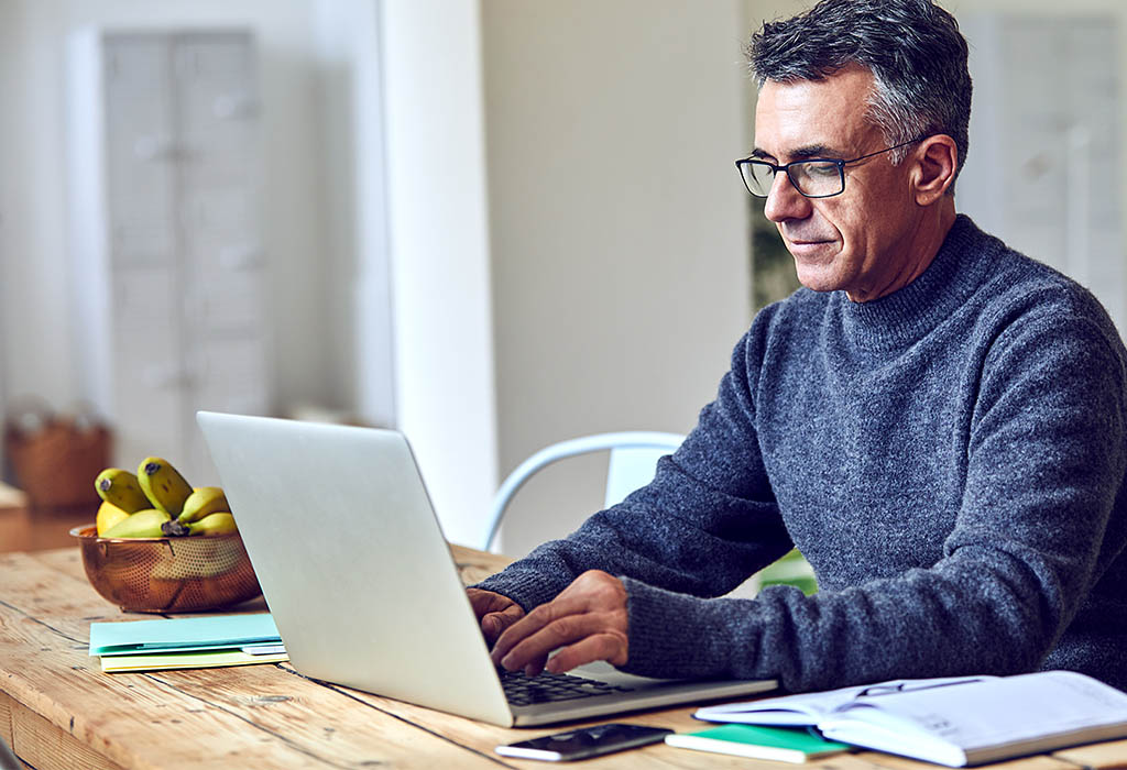 A middle aged man working on a laptop at home, he is wearing glasses, sweater, and collared shirt.