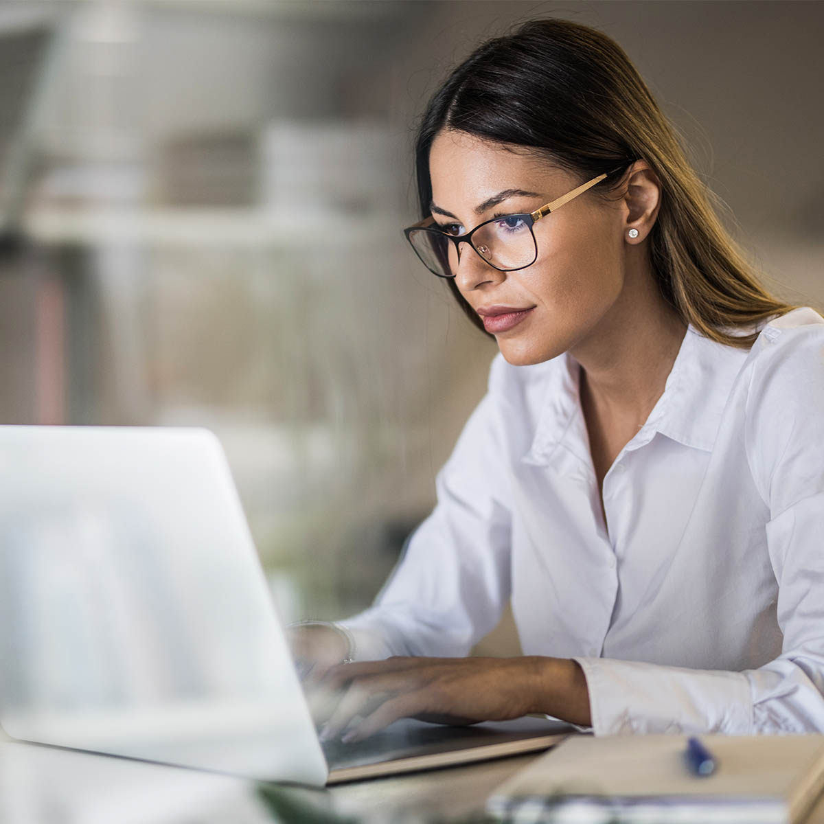 A mid-shot of a young business woman with glasses on working on a laptop in a quiet looking office.