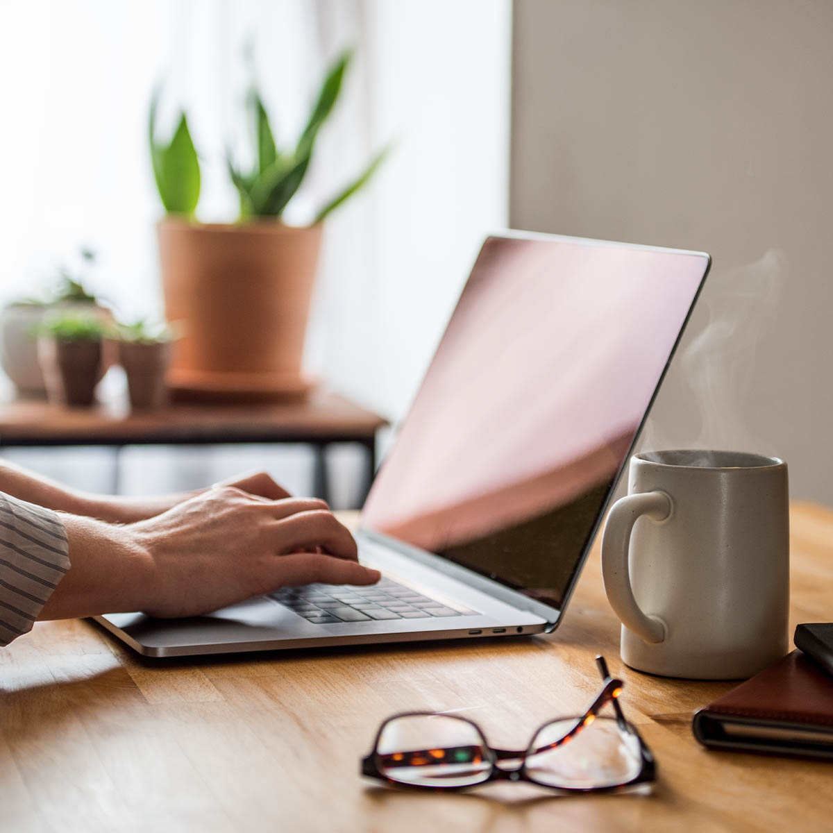 A womans hands typing a laptop at a wooden table with a cup and notebooks nearby.