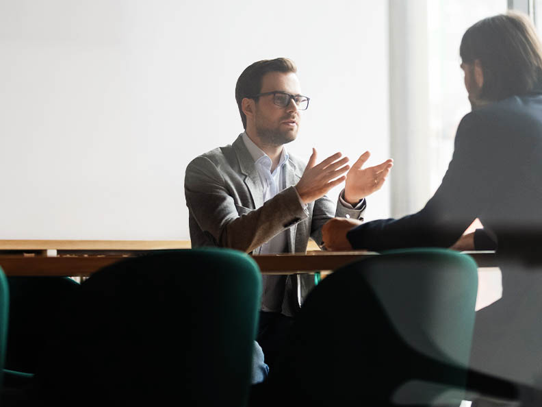 Mid shot image, shot through glass into a meeting room. A man with glasses and wearing a suit is gesturing and talking to a man across the table from him.