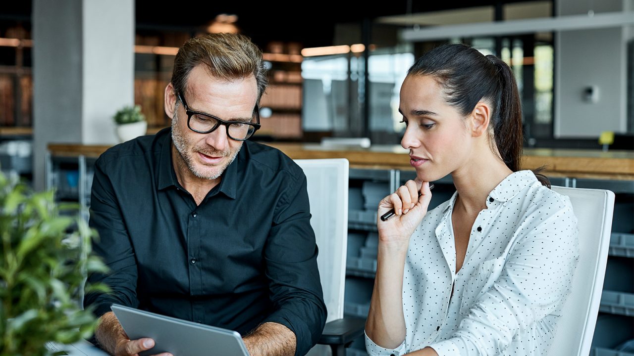 A male and female colleague looking at a tablet in a well lit office.