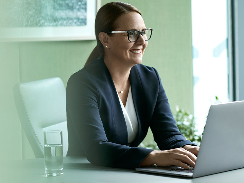 A businesswoman with glasses sitting at a desk working on laptop and smiling. Macquarie customer - Julia