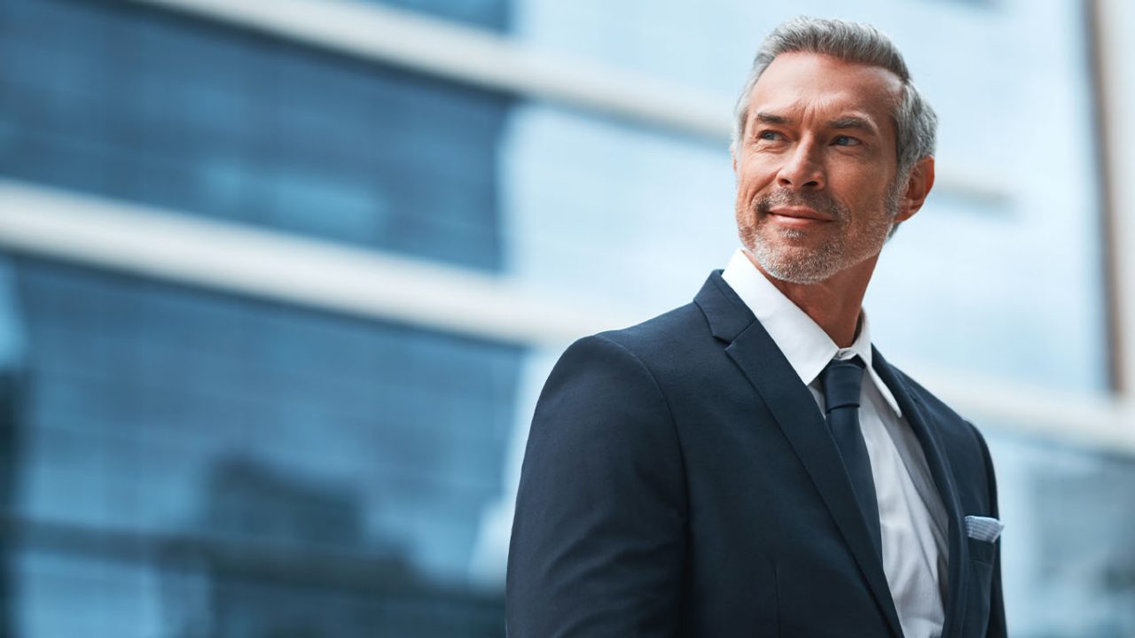 A middle aged well dressed business man looking off to the side, with his phone in his hands, standing in a city during the day.