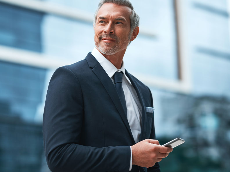 A middle aged well dressed business man looking off to the side, with his phone in his hands, standing in a city during the day.