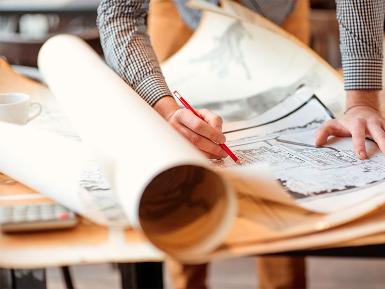 A person spreads out a blueprint and writes on it with pencil on a drafting table.