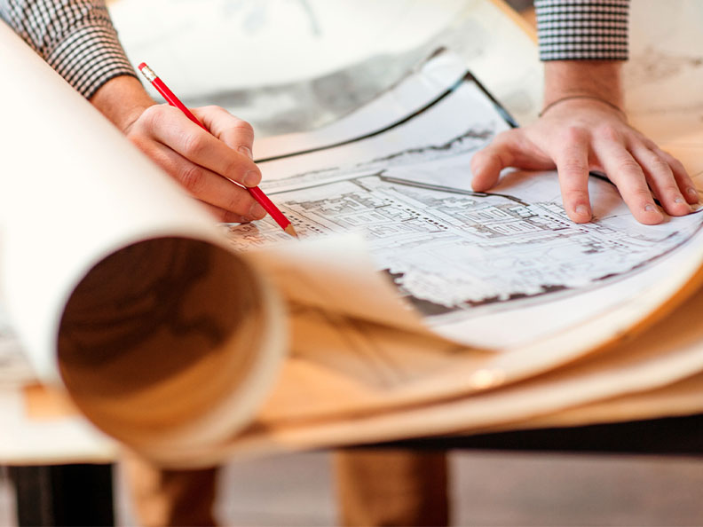 A person spreads out a blueprint and writes on it with pencil on a drafting table.