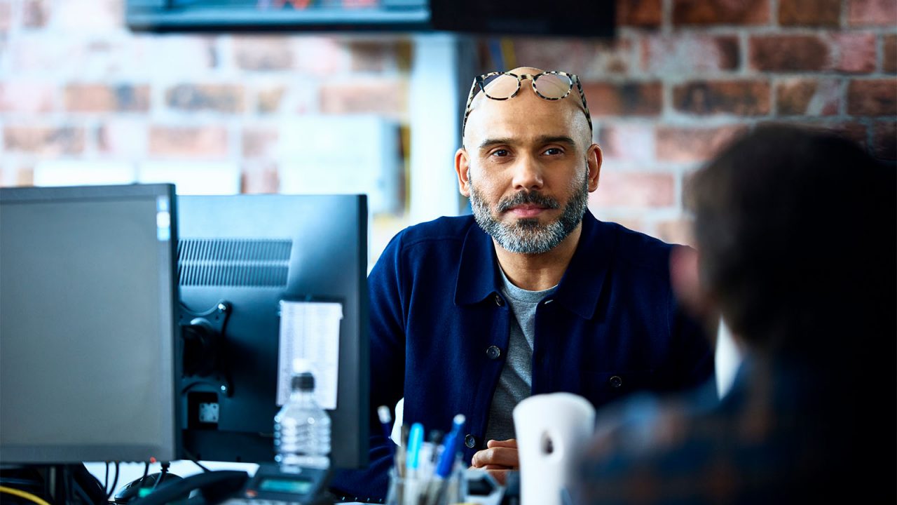 Two men talk across their desks with personal desktops in front of them in a modern brick office.