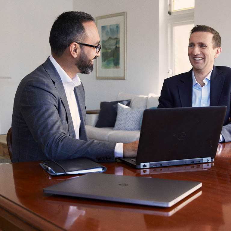 Two businessmen in suits sitting in a home lounge room, at a wooden table, looking at each other with a laptop