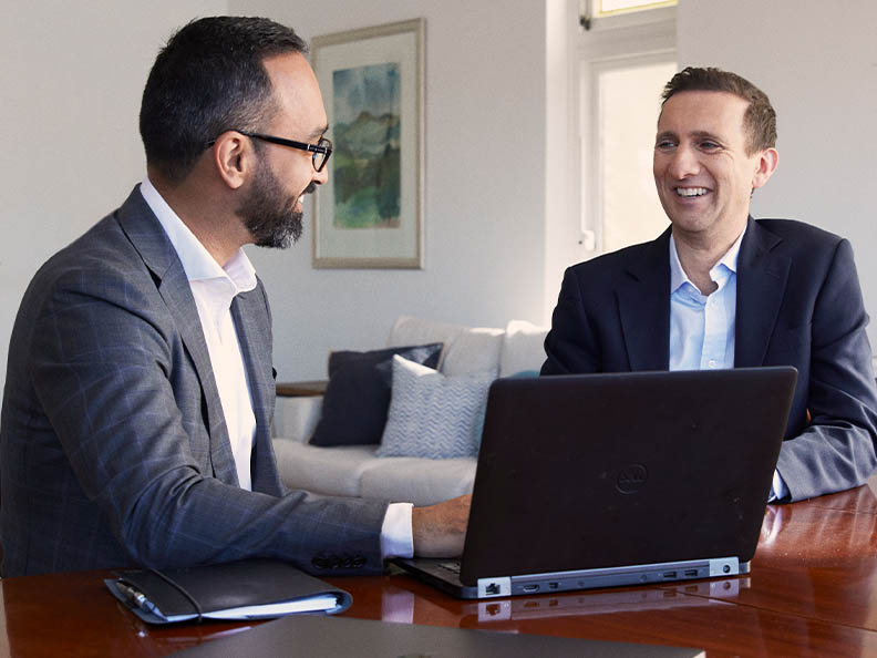 Two businessmen in suits sitting in a home lounge room, at a wooden table, looking at each other with a laptop