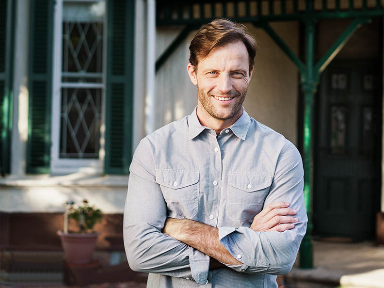 A photo of smiling man standing against house. Male is standing with arms crossed. He is wearing shirt.