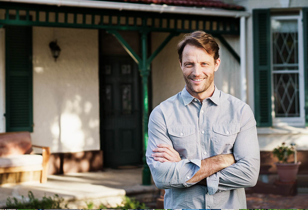 A photo of smiling man standing against house. Male is standing with arms crossed. He is wearing shirt.