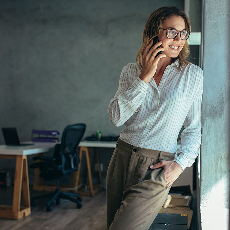 Businesswoman leaning against a wall, talking on cell phone and smiling. Female entrepreneur standing by a window and talking over mobile phone in office