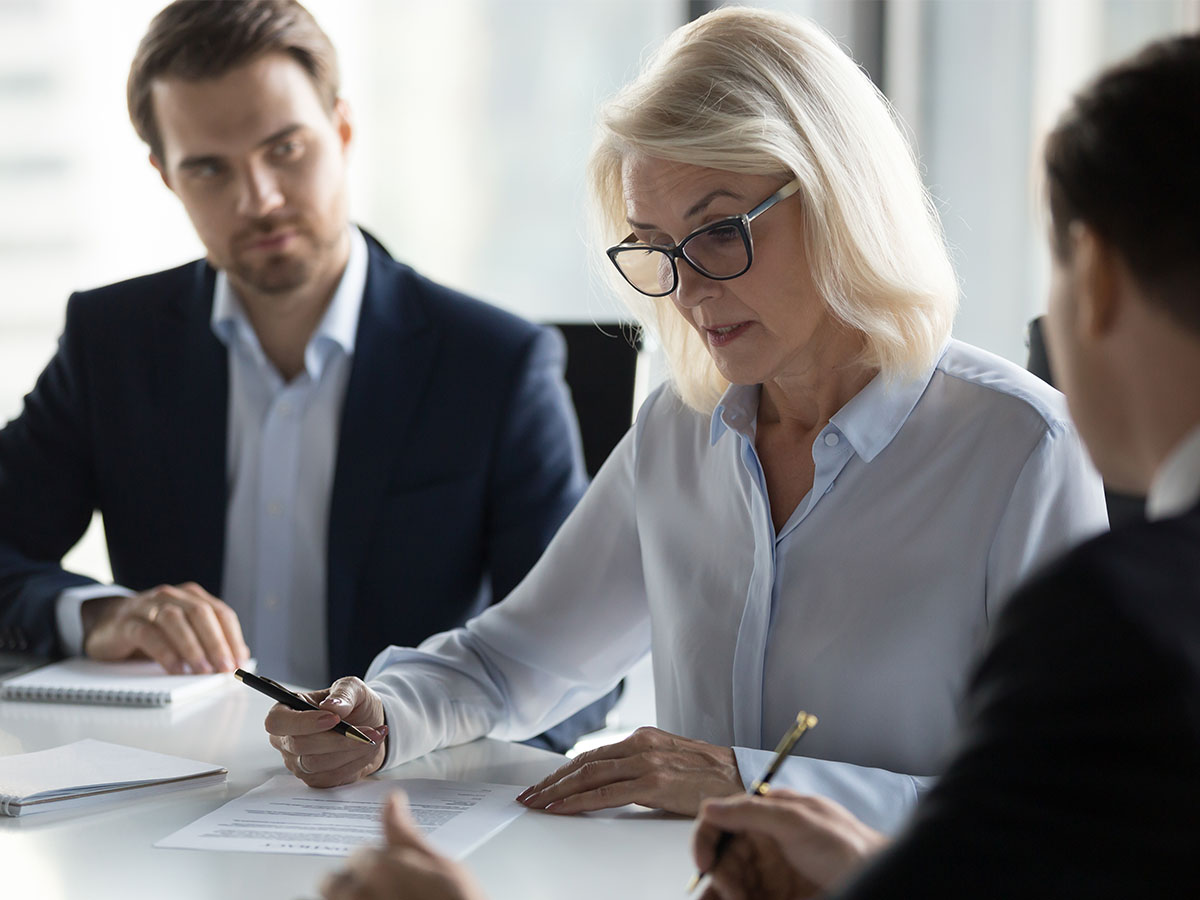 Businessmen sitting at desk headed by middle aged serious concentrated female in eyeglasses checking agreement document before signing it. Financial director ready affirm official paper with signature