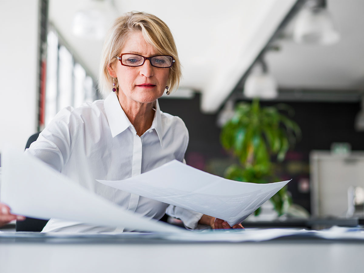 A mature aged business woman with glasses holding paper in each hand in a well lit office space