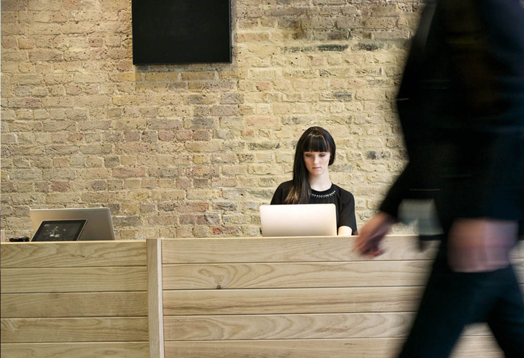 Dark haired receptionist working in an office with a person walking past out of focus.
