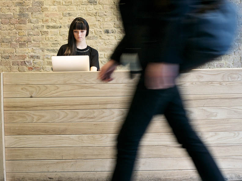Dark haired receptionist working in an office with a person walking past out of focus.