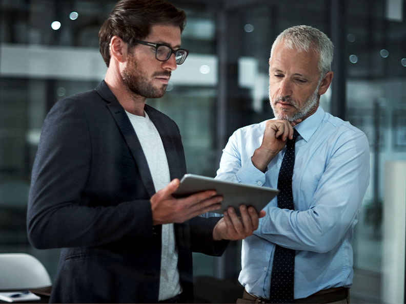 A mid shot of two business men looking at a smart tablet together in the office at night. One man is middle aged and the other is younger.