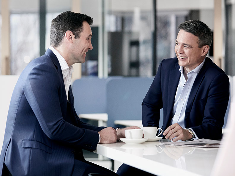 Two professionally dressed businessmen sitting and talking at a desk in a bright office over coffee