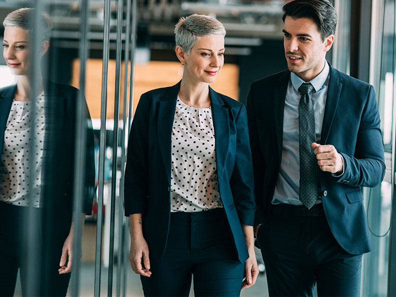Two colleagues dressed professionally in business attire walk through their office talking.