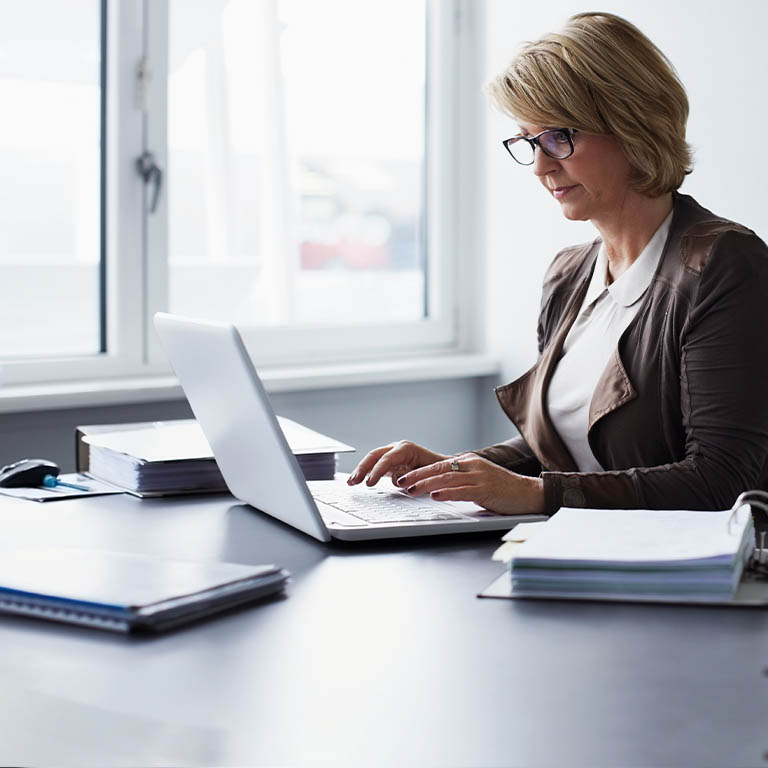 A middle aged woman with glasses sitting at a desk and looking at a laptop as she types