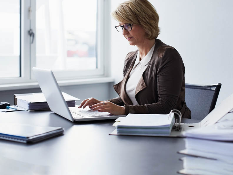 A middle aged woman with glasses sitting at a desk and looking at a laptop as she types