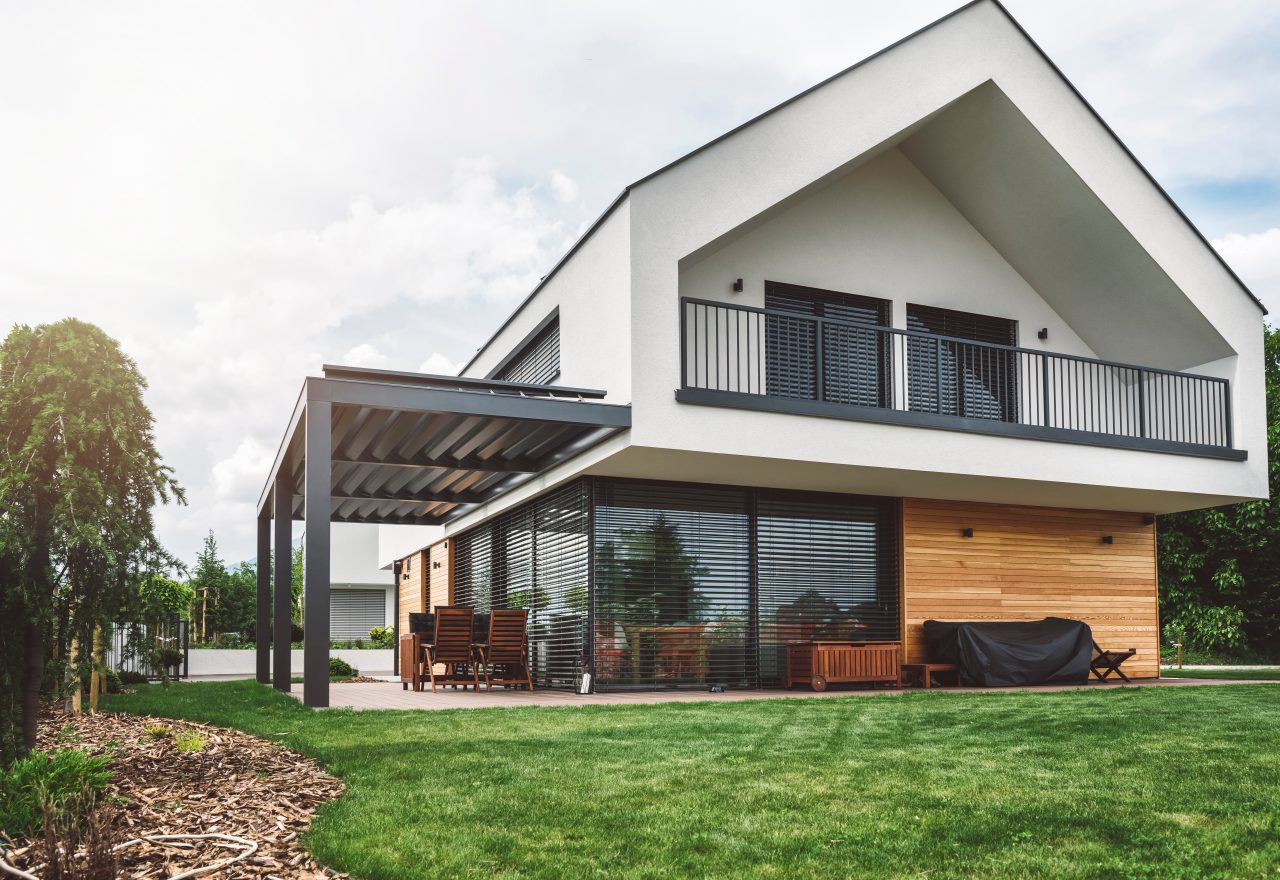 Modern backyard in the suburbs, big windows, wooden chairs and a modern picnic table, white facade of the modern building exterior, beauty of the nature and an amazing upper class residential district.