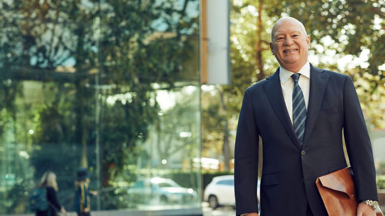 A businessman in a suit standing outside smiling as he holds some documents in his hand on a sunny day. Macquarie - Craig