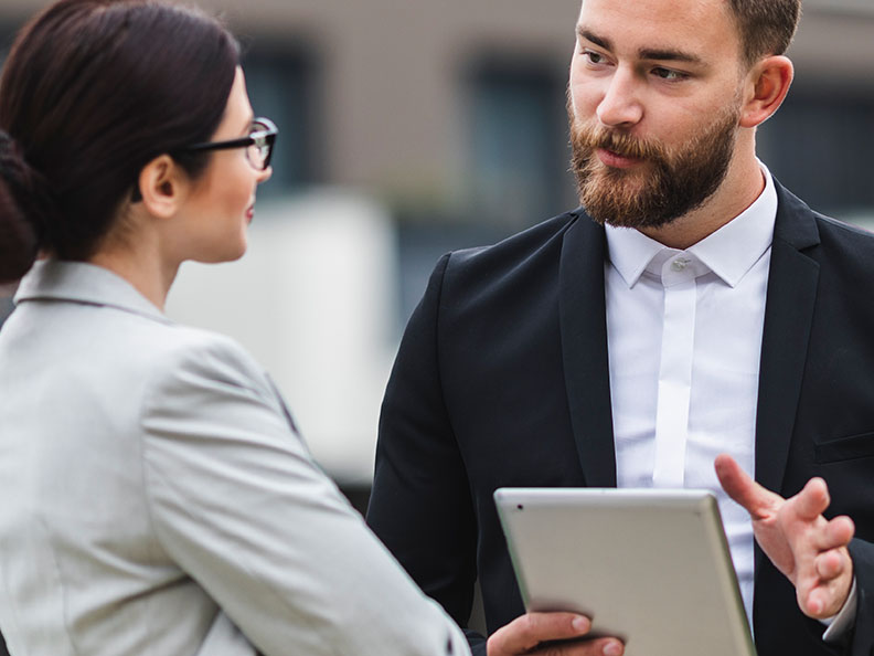 A man holding a tablet speaking to a professionally dressed woman outside.