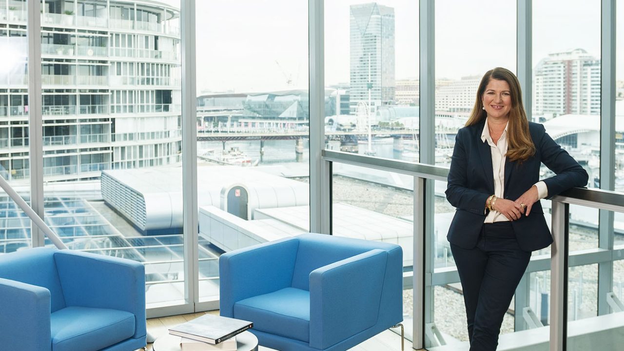 A professionally dressed woman in a brightly lit office building foyer leaning againt a railing