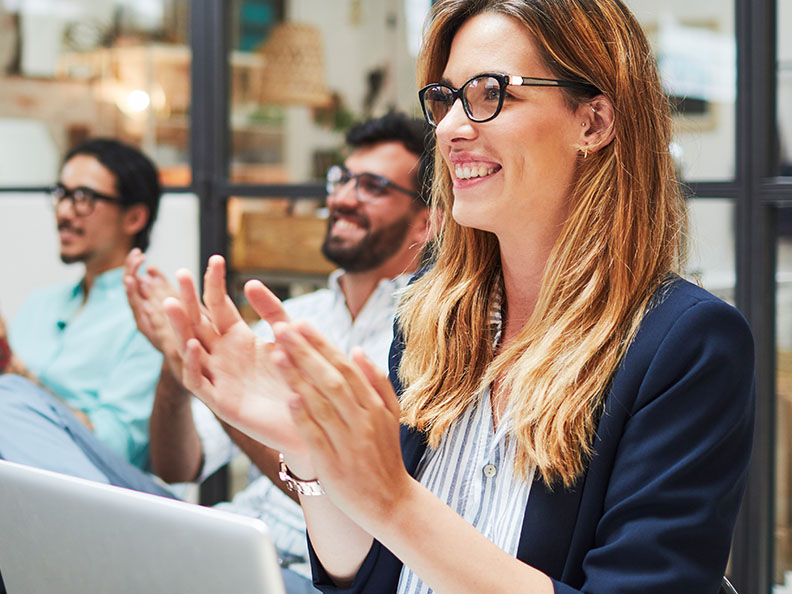 Business woman smiling and clapping with her peers during a conference style event in a meeting room. She has a laptop on her lap.