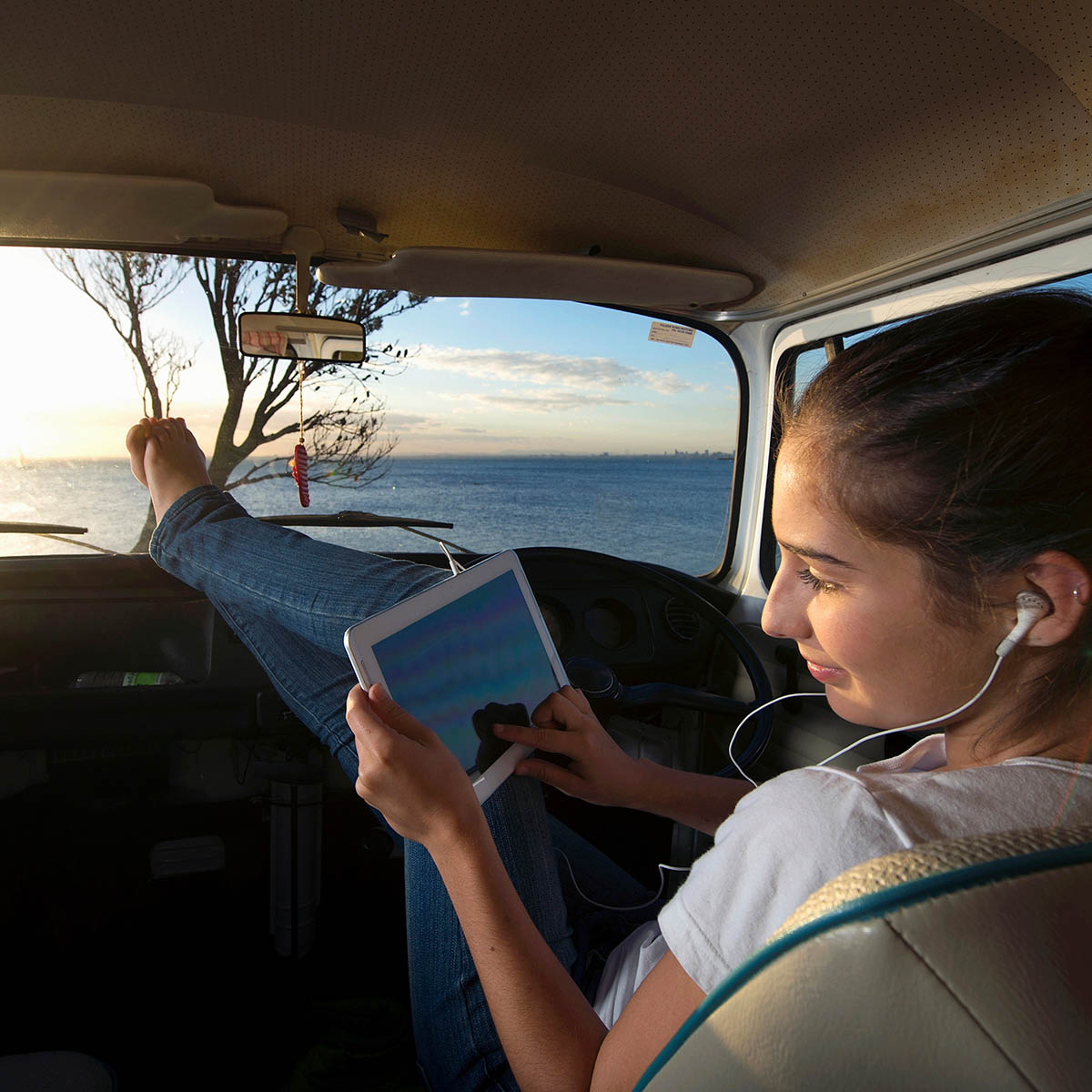 Girl with feet up in car interacting with tablet with ocean and sunset in the Background