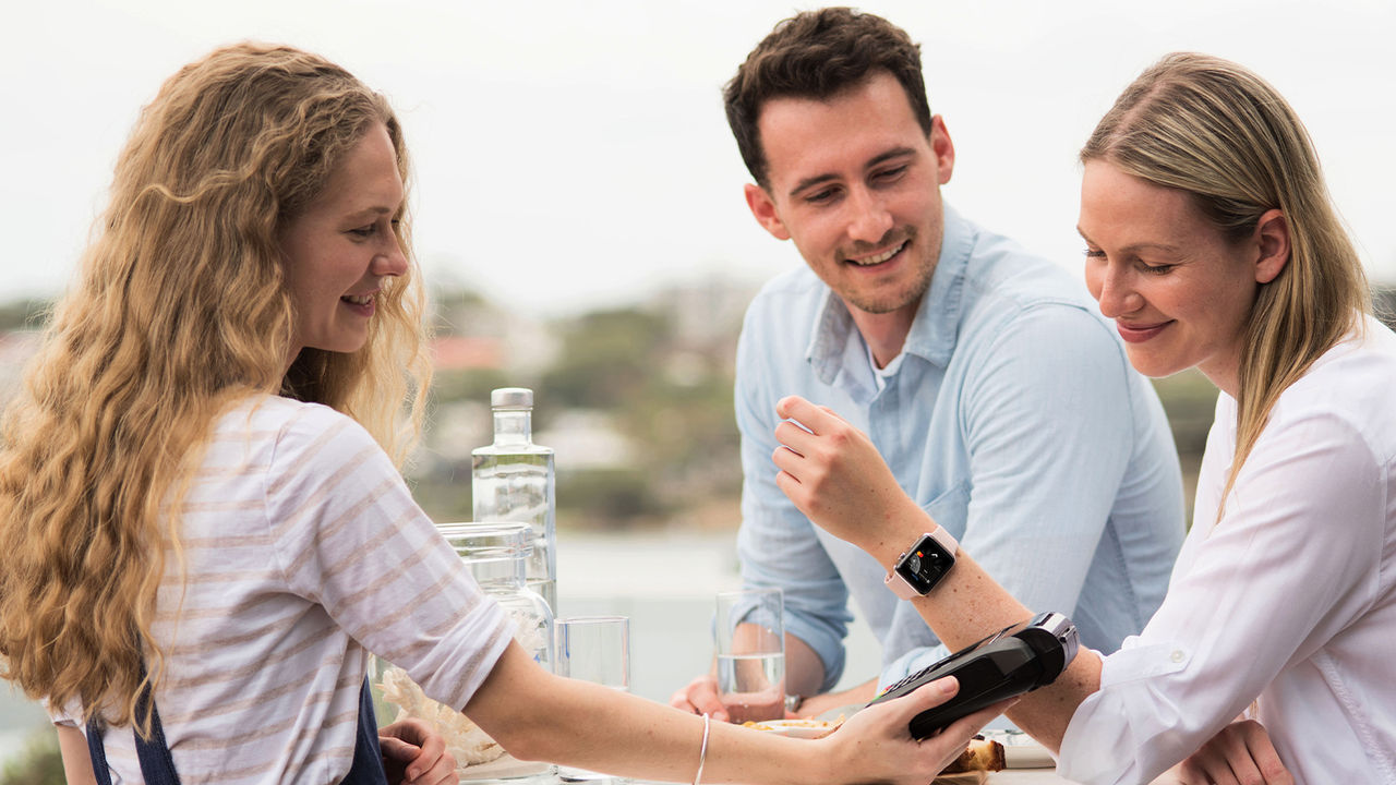 A woman pays with an Apple watch and a man sitting next to her smiling, outside. Macquarie - Apple Pay