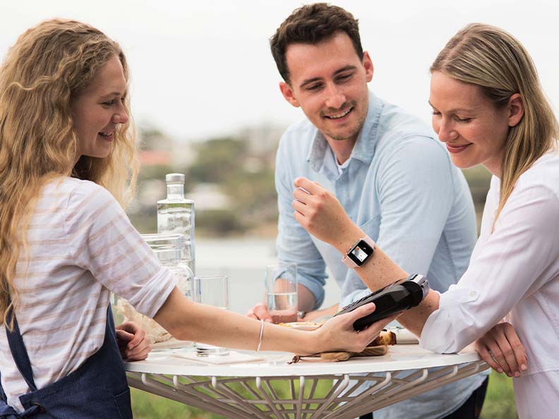 A woman pays with an Apple watch and a man sitting next to her smiling, outside. Macquarie - Apple Pay