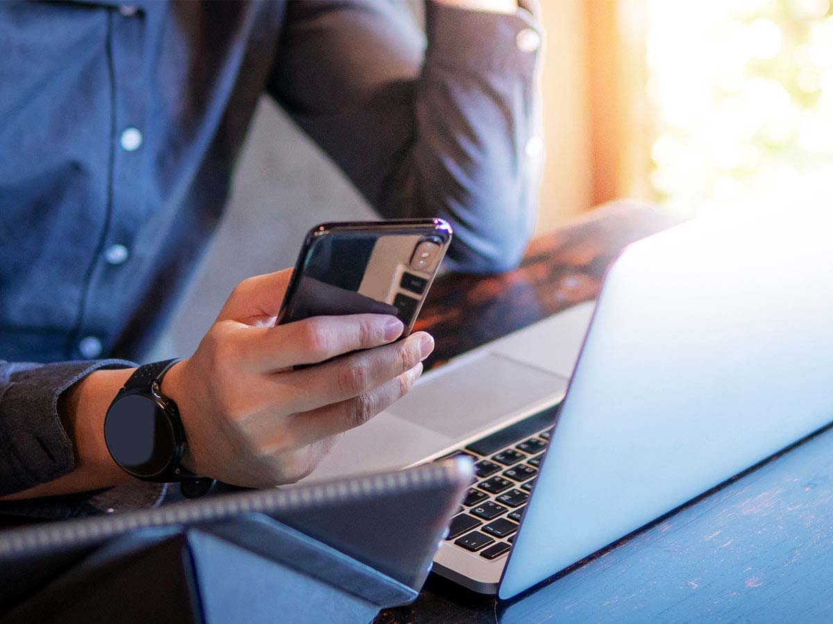 Hand holding a phone and wearing a smart watch with a laptop tablet in front of them