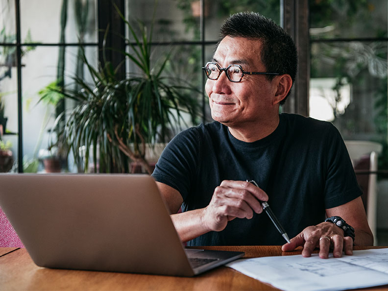 A close shot of a mature age Asian man holding a pen in a modern kitchen with a laptop in front of him with some paper documents.