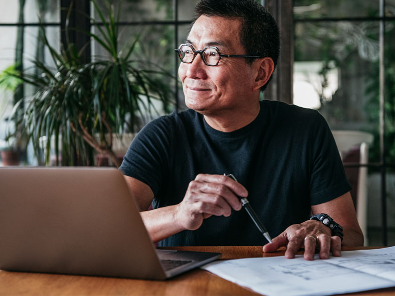 A close shot of a mature age Asian man holding a pen in a modern kitchen with a laptop in front of him with some paper documents.