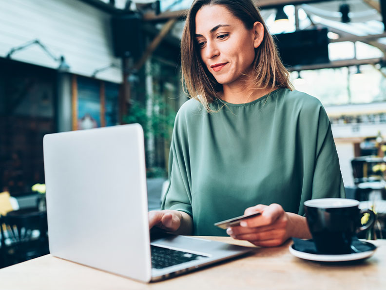 Casually dressed woman sititng in a cafe with a coffee, laptop open, and credit card in her hand