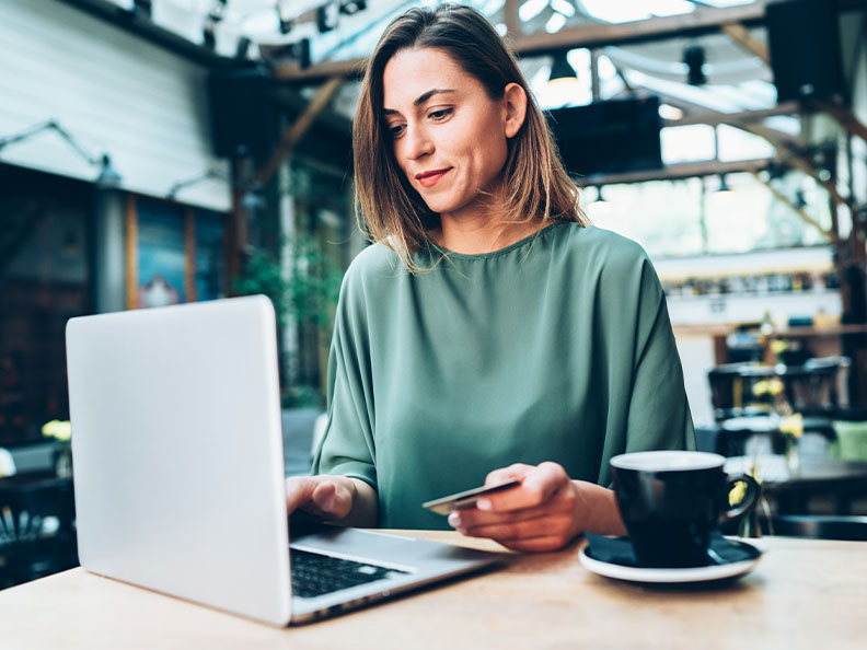 Casually dressed woman sititng in a cafe with a coffee, laptop open, and credit card in her hand