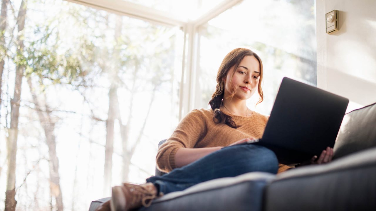 Young woman sitting casually on a couch looking at a laptop in a bright room
