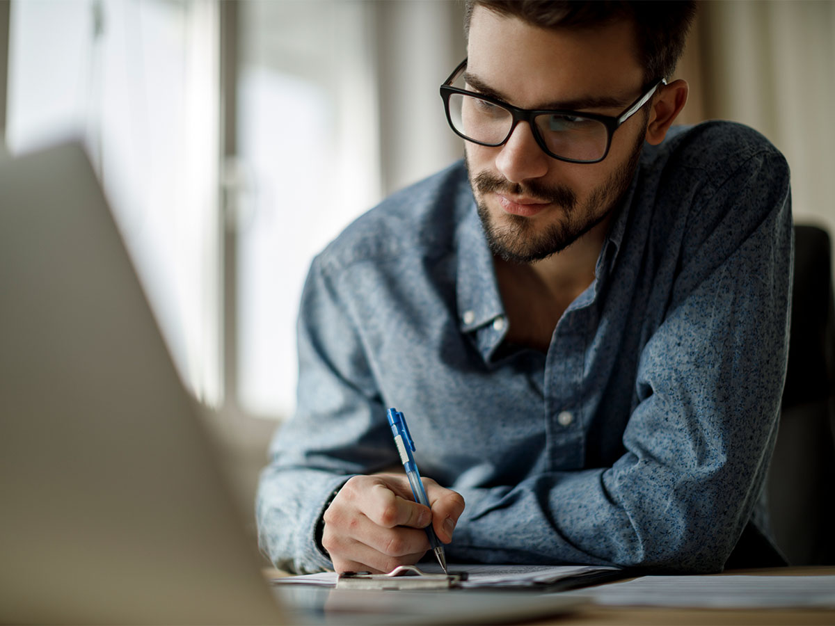 Medium/close up shot of a young man wearing glasses looking at a laptop and taking notes.