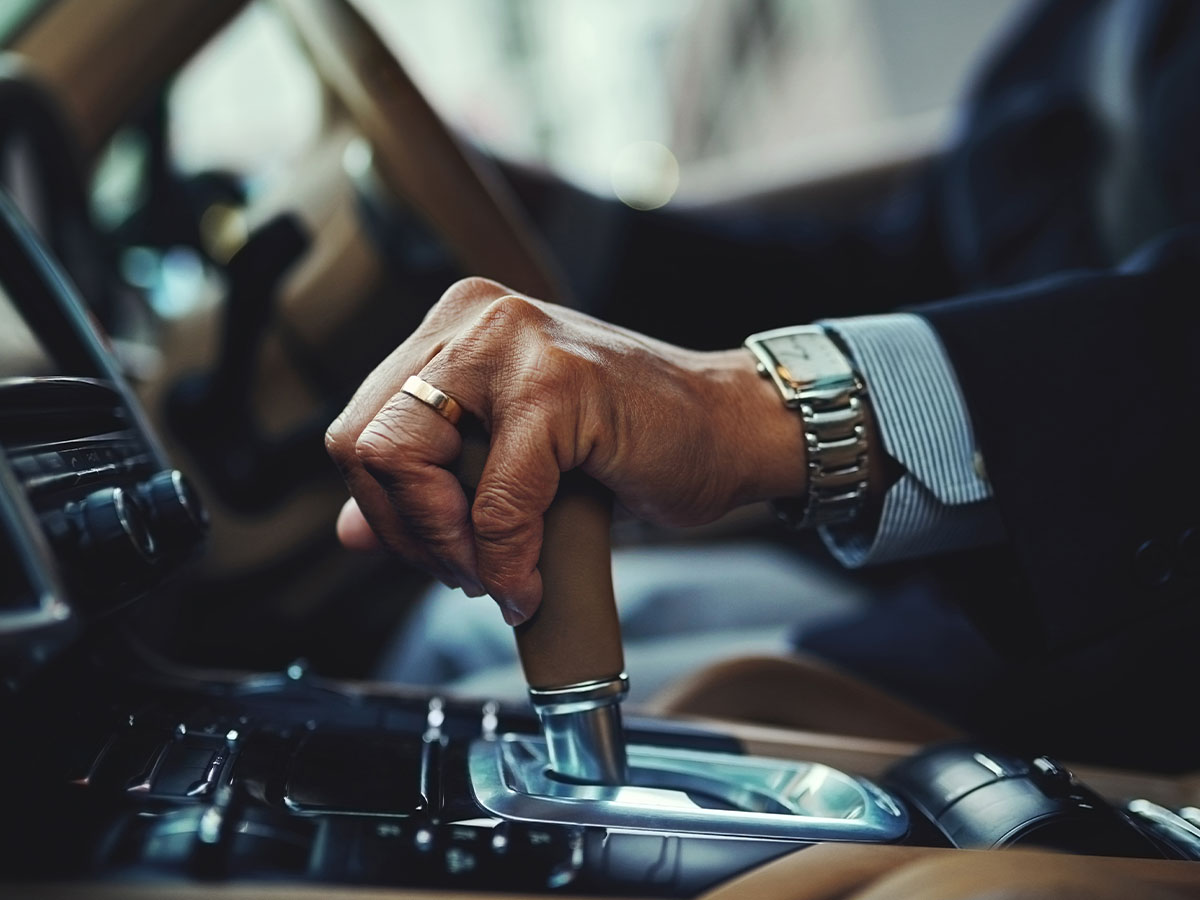 A person in a suit in a car with their hand with a watch and ring on holding onto the gear shifter of a car.