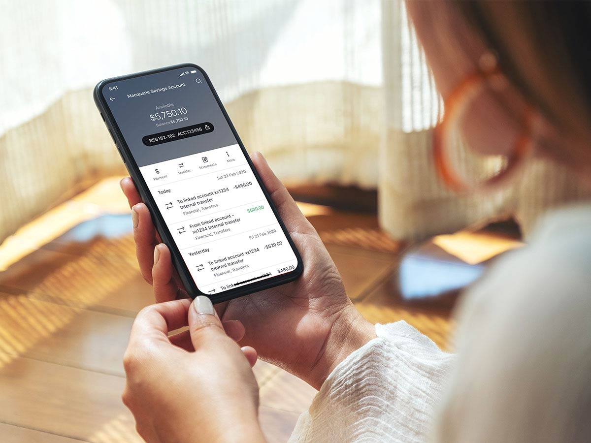 A woman holding a phone in a well lit room with the Macquaire bank app open on a savings screen.